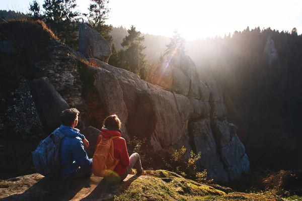 Два мандрівники сидять на краю скелі на лісистих пагорбах і хмарному небі на сході сонця. Чоловік і жінки вітають сонце . — стокове фото