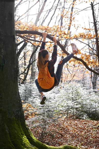 Joven mujer feliz colgando en el árbol en el bosque con mochila naranja. Divertida chica tonteando sobre un fondo de bosque . — Foto de Stock