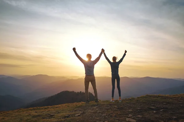 Silueta del hombre y la mujer de pie en el borde de la montaña y sosteniendo las manos en el cielo puesta del sol y el fondo de las montañas . —  Fotos de Stock