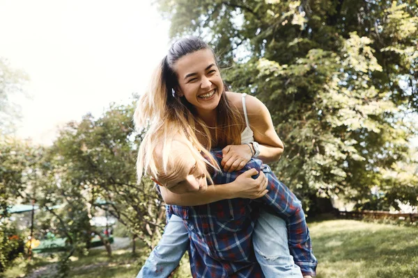 Mladý pár se baví a směje se v parku. Přítel s přítelkyní na zádech. — Stock fotografie
