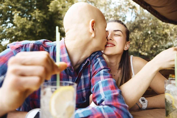 Молодая романтическая влюбленная пара проводит время в парке, сидит в летнем кафе и пьет холодный лимонад. Молодой человек нежно целует молодую женщину . — стоковое фото