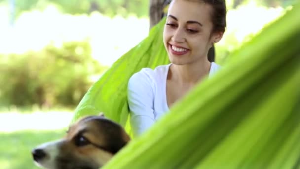 公園で明るい緑のハンモックに横たわっているかわいいウェールズコーギー犬と若いかわいい笑顔の女性 — ストック動画