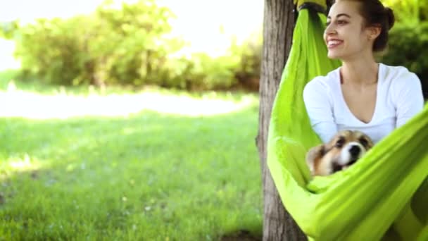 Молодая красивая улыбающаяся женщина с милой валлийской собакой Корги, лежащей в ярко-зеленом гамаке в парке — стоковое видео