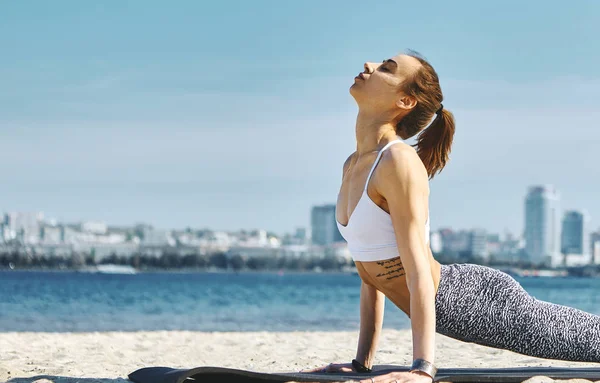 Primer plano retrato de mujer atlética joven delgada hace ejercicios de yoga y estiramiento en la playa de arena con fondo de ciudad. Vida sana, ejercicios matutinos, meditación . — Foto de Stock
