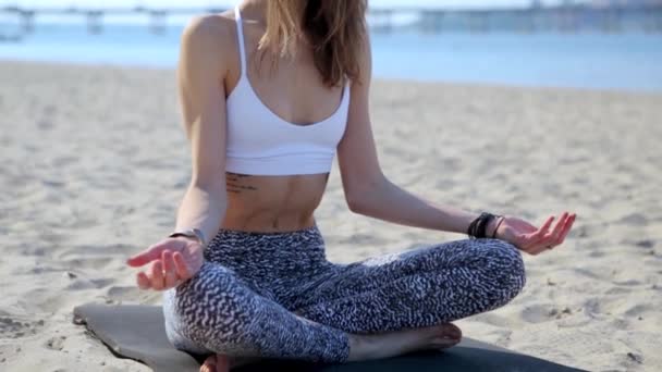 Portrait of a beautiful relaxed young woman sitting in lotus pose at the beach with sea and city background. womans hands in namaste murda pose — Stock Video