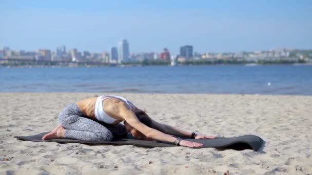 Молода струнка атлетична жінка робить вправи для йоги і тягнеться на піщаному пляжі з міським фоном. Здоровий спосіб життя, ранкові вправи, медитація . — стокове відео