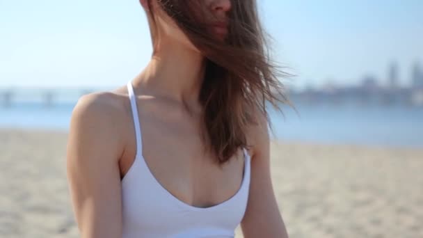 Portret van een mooie ontspannen jonge vrouw zittend in Lotus pose op het strand met zee en stad achtergrond. Dames handen in Namaste Murda pose — Stockvideo