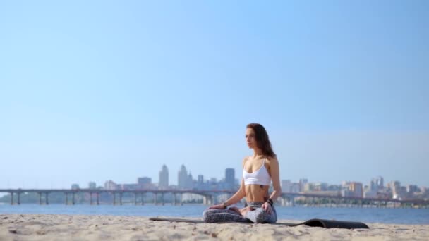 Портрет красивой расслабленной молодой женщины, сидящей в позе лотоса на пляже на морском и городском фоне. женщины руки в позе namaste murda — стоковое видео