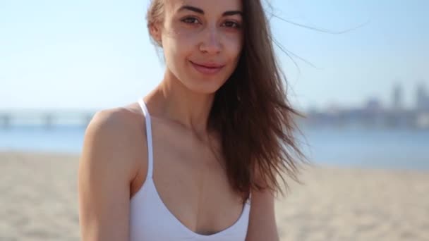 Feliz mujer de raza mixta alegre de etnia caucásica asiática sentada en la playa y mirando a la cámara, viento soplando el pelo de las mujeres — Vídeo de stock