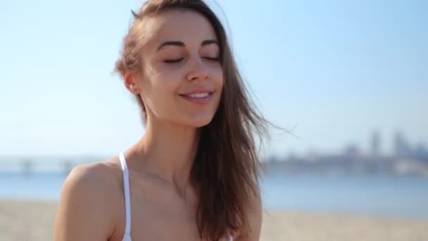 Feliz mujer de raza mixta alegre de etnia caucásica asiática sentada en la playa y mirando a la cámara, viento soplando el pelo de las mujeres — Vídeo de stock