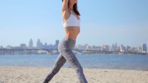 Mujer practicando yoga-asanas uotdoors en la playa de arena. joven mujer atlética delgada de pie en yoga asana con las manos levantadas sobre la ciudad y el fondo azul del cielo . — Vídeos de Stock