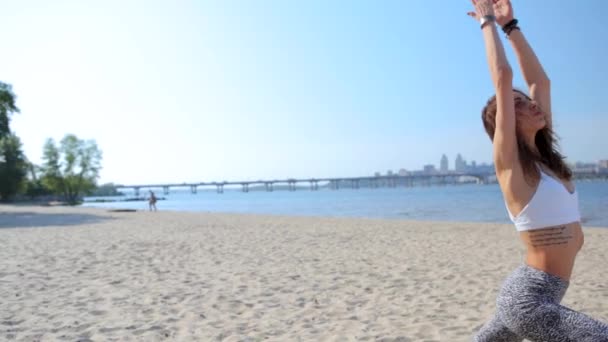 Donna che pratica l'esecuzione di yoga-asana uotdoors sulla spiaggia di sabbia. giovane donna atletica sottile in piedi in asana yoga con le mani sollevate sulla città e sfondo cielo blu . — Video Stock