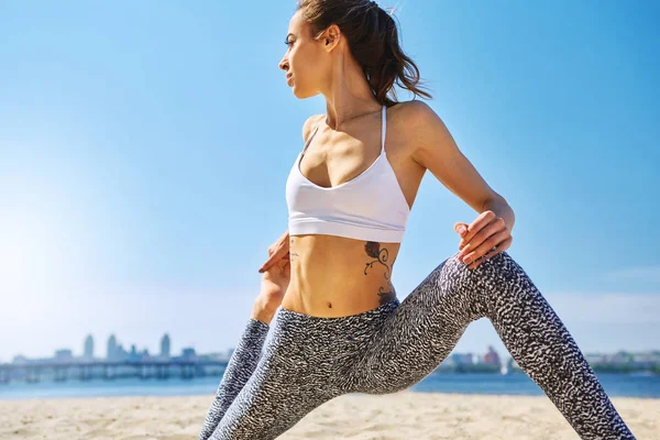 Lähikuva muotokuva nuori hoikka urheilullinen nainen tekee jooga harjoituksia ja venyttely hiekkarannalla kaupungin tausta. Terveellinen elämäntapa, aamuharjoitukset, meditaatio . — kuvapankkivalokuva