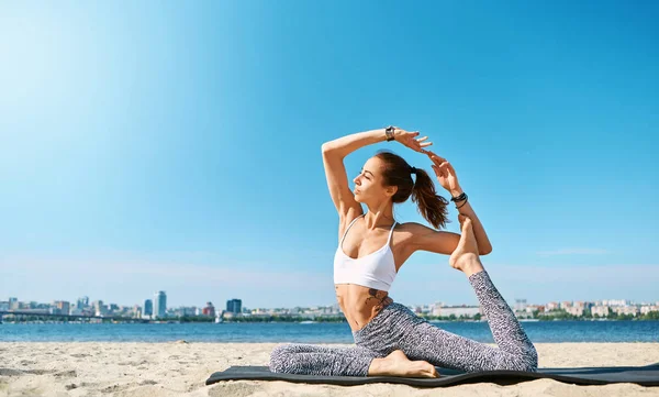 Joven mujer atlética delgada hace ejercicios de yoga y estiramiento en la playa de arena con fondo de ciudad. Vida sana, ejercicios matutinos, meditación . — Foto de Stock