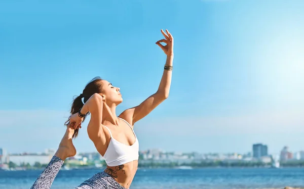 Ung, slank, atletisk kvinne gjør yogatrening og strekker seg på sandstranden med bybakgrunn mot blå himmel og hav. Sunn livsstil, morgenøvelser, meditasjon . stockbilde