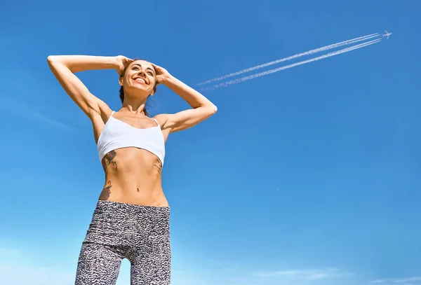 Onnellinen hymyilevä urheilu nainen ruskettunut istuvuus kehon ja tatuointi poseeraa sininen taivas lentokone polkuja . — kuvapankkivalokuva