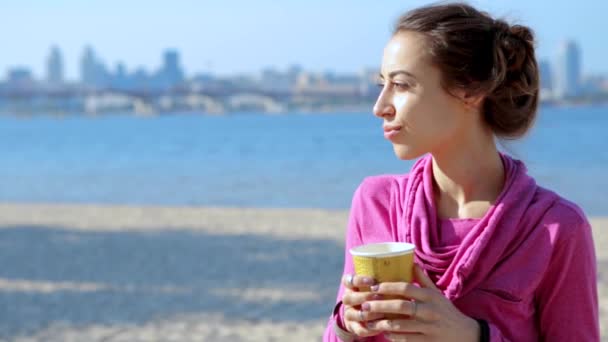 화창한 아침에 도시 모래 해변에서 종이 컵에서 커피를 마시는 분홍색 스포츠웨어 후드에 아름다운 젊은 운동 여성의 초상화. — 비디오