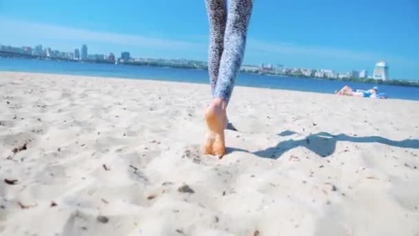 Vista posterior inferior de la hermosa mujer atlética delgada joven en leggins caminando en la playa de arena en la mañana soleada . — Vídeos de Stock