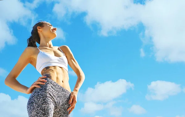 Onnellinen hymyilevä urheilu nainen ruskettunut sovi kehon ja tatuointi poseeraa sininen taivas pilvet, pohja näkymä . — kuvapankkivalokuva