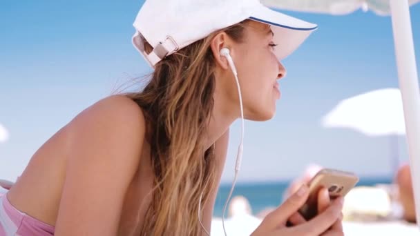 Beautiful relaxed slim sexy tanned woman in bikini and white cap with navy blue Union Jack British flag is lying on the beach on a white sand, relaxing and listen music from the phone with small Stock Footage