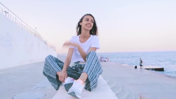 Krásná mladá žena v lehkých letních šatech, která za úsvitu sedí na molu. Mladá Kavkazská samice sleduje nádherný výhled na západ slunce. — Stock video