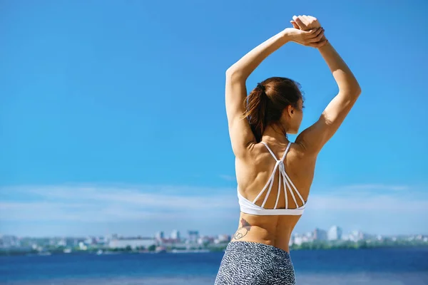 Attraktiv sportskvinne med solbrent kropp som poserer med hevet arm på stranden med bybakgrunn mot blå himmel og hav. bakover visning . – stockfoto