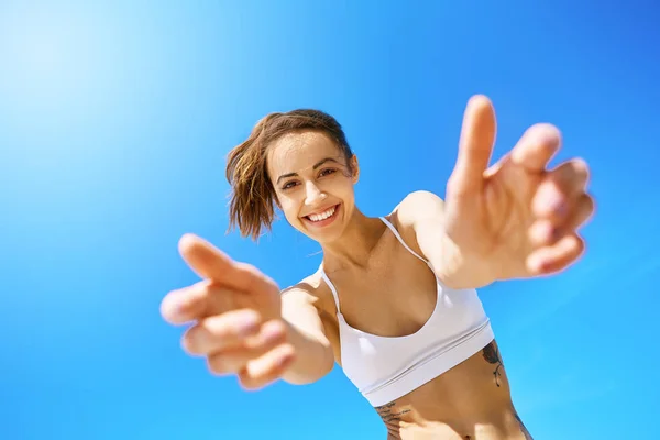 First person view of beautiful happy smiling woman with outstretched hands to the camera on blue sky background. Young smiling fitness woman having fun and laughing against blue clear sky. — Stock Photo, Image