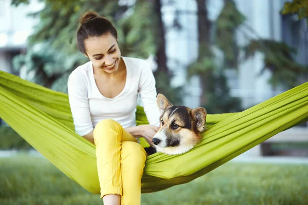 Smiling young woman in green hammock with cute dog Welsh Corgi in a park outdoors. Beautiful happy female in white shirt enjoying good day and petting her dog. Focus on the dog. — Stock Photo, Image