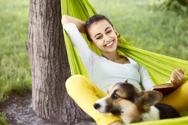 Leende ung kvinna i grön hängmatta med rolig hund walesiska Corgi i en Park utomhus. Vacker lycklig kvinna i vit skjorta njuter bra dag och läsa en bok. — Stockfoto