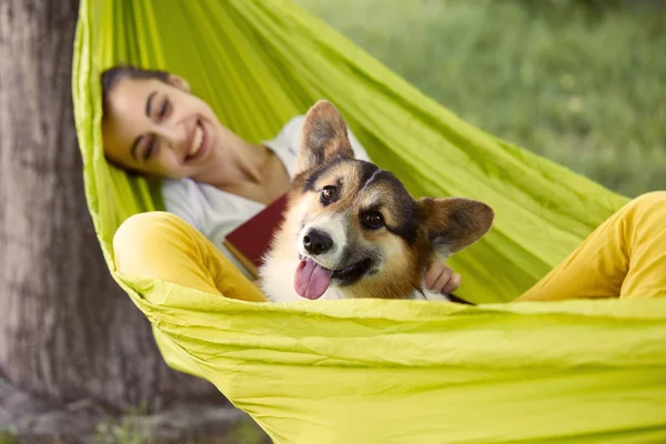 Smiling young woman resting in green hammock with cute dog Welsh Corgi in a park outdoors. Beautiful happy female in white shirt enjoying good day and looking on pet. Focus on the dog. — Stock Photo, Image