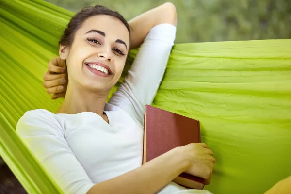 Portrait of a smiling young woman lying in green hammock with book in park. — Stock Photo, Image