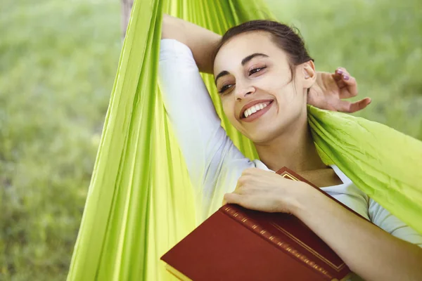 Portrait d'une jeune femme souriante couchée dans un hamac vert avec livre dans le parc . — Photo