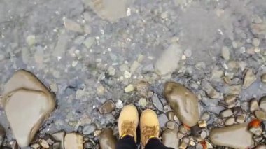 çakıl sahil ile dağ nehri yakınında sarı çizmeler kadın bacaklarınıilk kişi görünümü. kadın elini suya sokar ve yıkar. Pov. Dağ deresi berrak ve şeffaf su