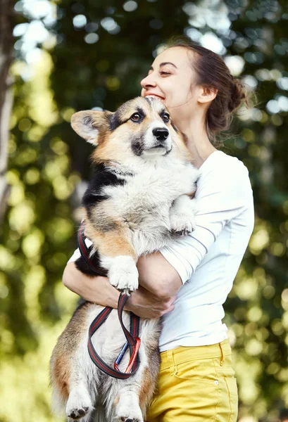 Glimlachende jonge vrouw die schattige hond Welsh Corgi in een park buitenshuis houdt. Focus op de hond. — Stockfoto