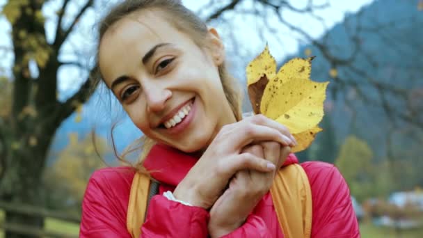 Красива усміхнена жінка в червоній куртці, що стоїть на відкритому повітрі, тримає кілька сухих жовтих листя в руках і грає з ними, грайливо ховається за листям і заглядає — стокове відео