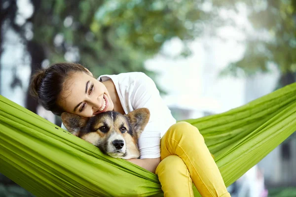 Young woman in green hammock with cute dog Welsh Corgi in a park outdoors. Beautiful happy female in white shirt enjoying good day and petting her dog. Focus on the dog. — Stock Photo, Image