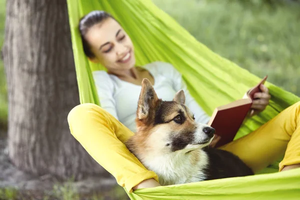 Leende ung kvinna i grön hängmatta med rolig hund walesiska Corgi i en Park utomhus. Vacker lycklig kvinna i vit skjorta njuter bra dag och läsa en bok. — Stockfoto