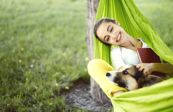 Jeune femme souriante dans un hamac vert avec chien mignon Welsh Corgi dans un parc à l'extérieur. Belle femme heureuse en chemise blanche profitant d'une bonne journée et embrassant un animal de compagnie. Focus sur le chien . — Photo