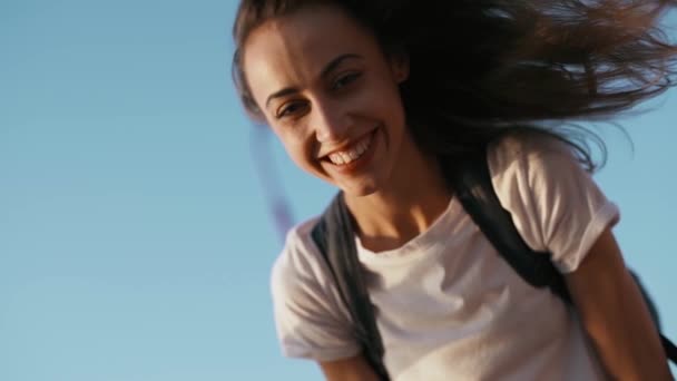 白いTシャツを着た幸せな笑顔の若いかわいい女性は、青空の背景に橋の上に立って、笑い、平和な夕日を楽しみ、カメラを見ています. — ストック動画