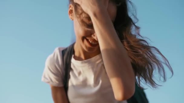 Joven mujer bastante alegre en camiseta blanca está de pie sobre el fondo del cielo azul, endereza el cabello a mano y mirando a la cámara. calma y relajación . — Vídeo de stock