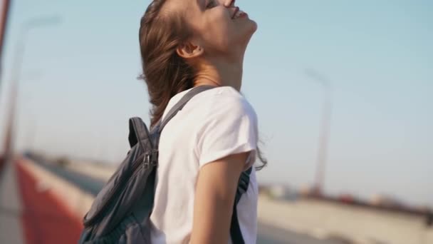 Jong volwassen vrouwelijk model poseren buitenshuis. Gelukkig ontspannen mooie vrouw in wit t-shirt staat op de brug op blauwe hemel achtergrond, genieten van rustige zonsondergang en wind, kijken naar camera. — Stockvideo