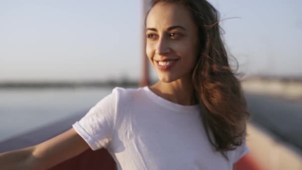 Jonge gelukkige vrouw kijkt 's avonds zonsondergang op zee. mooi meisje staat op de brug, genietend van het uitzicht op de zonsondergang en lichte frisse wind — Stockvideo