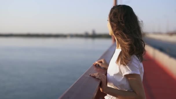 Jovem mulher feliz olhando à noite pôr do sol no mar. menina bonita está de pé na ponte, apreciando a vista do pôr do sol e luz do vento fresco — Vídeo de Stock