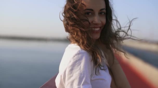 Молодая счастливая женщина смотрит на вечерний закат в море. симпатичная девушка стоит на мосту, наслаждаясь закатным видом и легким свежим ветром — стоковое видео