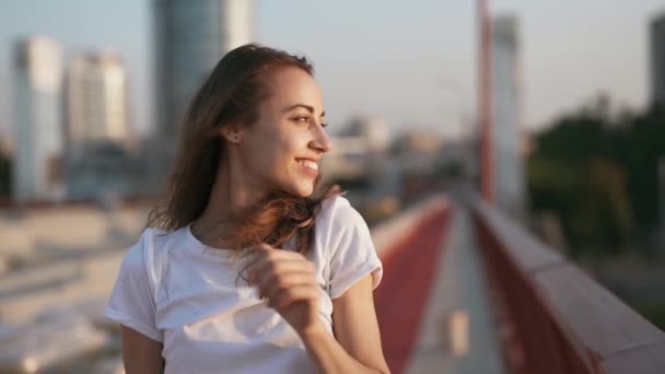 Щаслива усміхнена молода красива жінка в білій футболці стоїть на мосту на фоні блакитного неба, рука вирівнює волосся, насолоджуючись спокійним заходом сонця і дивлячись на камеру . — стокове відео