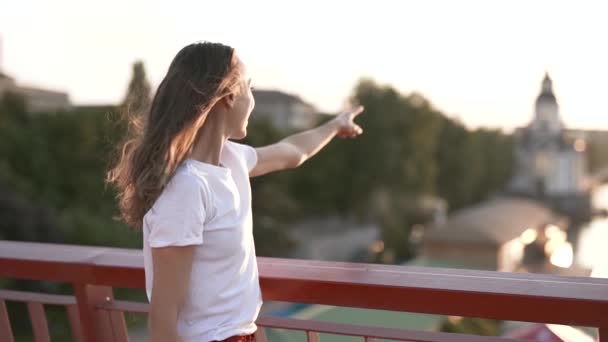 Feliz mujer hermosa joven y sorprendida en camiseta blanca está de pie en el puente sobre el fondo de la ciudad, alegremente mostrando delante de la mano, disfrutando de la puesta de sol pacífica y mirando hacia el futuro . — Vídeos de Stock