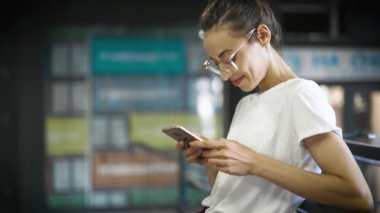 beyaz t-shirt ve şeffaf gözlük genç gülümseyen şık güzel kadın açık havada akıllı telefon kullanıyor. kadın bir şey arıyor, sosyal ağları scroling, sohbet ve mesajlaşma.