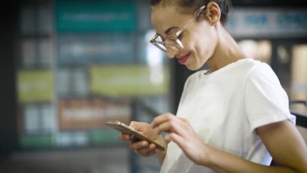 Ung leende snygg vacker kvinna i vit t-shirt och transparenta glasögon använder smartphone utomhus. kvinna söker något, scroling sociala nätverk, chating och textning. — Stockvideo