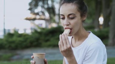 beyaz t-shirt çekici genç kadın yaz akşamı parkta açık havada oturuyor ve kağıt fincan kahve içme ve waffle yiyor. kadın bir ısırık yapar ve yemek bir tat sahiptir.