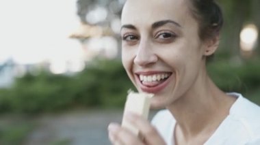 beyaz t-shirt closeup çekici genç kadın yaz akşamı parkta açık havada oturuyor ve waffle yiyor. kadın kamera için bir çerez önerir ve bir ısırık yapar ve yemek bir tat sahiptir.
